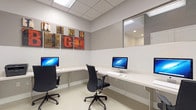 This single-floor space functions as a dedicated business center, designed with a clean, modern aesthetic that prioritizes functionality and productivity. The room features a continuous built-in white desk that wraps along two walls in an L-shaped configuration, providing ample workspace for multiple users simultaneously. Two desktop computers are stationed along the desk, each paired with an ergonomic black office chair that offers comfortable, adjustable seating.

The room's interior is finished with neutral gray and white walls, complemented by light-toned tile flooring that contributes to the bright, professional atmosphere. Overhead, a suspended ceiling with recessed panel lighting ensures consistent, even illumination throughout the workspace. A large interior window is positioned along one wall, allowing borrowed light from an adjacent space and adding a sense of openness to the otherwise compact room.

A standout decorative element is the bold "Think Big" typographic wall art displayed prominently above the desk. Rendered in a mixed-media, distressed style with warm red and earthy tones, it adds visual interest and an inspirational tone to the environment. The overall ambiance is tidy, efficient, and purposefully designed to support focused work in a shared setting.
