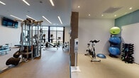 Located at 7323 W 38th Ave in Wheat Ridge, Colorado, this well-appointed fitness center offers a comprehensive workout environment across a single floor. The space is divided into distinct functional zones, each designed to support a variety of training styles and fitness goals.

The main strength and cardio area is bathed in natural light from floor-to-ceiling windows that frame outdoor views, creating an open and energizing atmosphere. This section houses a full rack of dumbbells, multiple cable and multi-use strength machines, adjustable benches, treadmills, ellipticals, and a wall-mounted flat-screen TV. Large mirrors amplify the sense of space and allow users to monitor their form. Recessed and linear LED lighting supplements the abundant daylight.

A dedicated functional training zone features a power rack with a training station, a water rowing machine, and ample open floor space for dynamic movement. An adjacent studio area is equipped with stationary cycling bikes, stability balls, a BOSU trainer, and a selection of medicine balls and smaller dumbbells, all stored on organized racks. This section is finished with accent wall color and recessed lighting, providing a distinct, focused environment.

Throughout the facility, a geometric wood shelving unit offers convenient storage for towels and accessories. Sanitation stations are positioned at entry points, and durable, easy-to-clean flooring runs throughout the space.