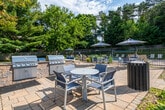 This single-story community pool facility offers a well-appointed outdoor recreational experience surrounded by mature trees and manicured greenery. The grounds are anchored by a swimming pool enclosed within a sleek black metal fence, with a generously sized stone paver deck lined with lounge chairs for relaxing in the sun.

Adjacent to the pool area, a spacious patio extends the outdoor living space with a stylish seating arrangement centered around a fire pit table, providing an inviting spot for gathering. A built-in outdoor kitchen station featuring a stainless steel gas grill with a granite countertop and stone base adds excellent amenity for outdoor entertaining.

A dedicated pool house structure clad in dark green vertical board siding serves as the facility's entry point. Inside, a tidy foyer with light blue beadboard walls, tile flooring, and white panel doors provides access to the restroom facilities and utility areas. Pool rules and operational hours are clearly posted on the exterior wall of the pool house, indicating supervised usage guidelines for residents.

Stone paver pathways connect the pool house to the surrounding pool deck, creating a cohesive and well-designed flow throughout the facility. The entire space benefits from abundant natural light and a backdrop of lush, towering trees, lending the area a serene and private atmosphere ideal for outdoor recreation and relaxation.