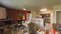 This single-floor residence showcases an open-concept layout that seamlessly connects the living area and kitchen, creating a spacious and functional environment. The living room is anchored by a striking terra cotta accent wall, which adds warmth and visual depth to the space. A ceiling fan with an integrated light fixture is centrally mounted overhead, providing both comfort and illumination. The room accommodates a comfortable mix of seating, including a tufted leather chair and a cushioned sofa adorned with patterned throw pillows, all arranged atop a decorative area rug with bold, colorful motifs. A circular mirror cluster serves as a prominent wall feature, enhancing the room's character.

The kitchen is accessible via a raised breakfast bar fitted with rustic wooden bar stools, creating a natural transition between the two spaces. The kitchen is equipped with stainless steel appliances, including a refrigerator and microwave, complemented by rich dark wood cabinetry. Light-toned countertops and neutral wall colors balance the darker elements throughout. The overall ambiance of the space is warm and inviting, with earthy tones and carefully chosen décor contributing to a cohesive, homey aesthetic.