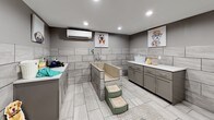 This purpose-built pet wash facility is thoughtfully designed to provide a clean, functional, and welcoming environment for pet owners and their animals. The space features large-format, light grey stone-look tile covering both the floors and walls halfway up, creating a cohesive, easy-to-clean aesthetic throughout. Recessed lighting illuminates the interior evenly, contributing to a bright and practical atmosphere.

At the heart of the space are built-in pet bathing tubs, constructed with matching tile and equipped with mounted faucets and handheld spray attachments. Convenient pet steps sit alongside the tubs, allowing easy access for animals of all sizes. Grey cabinetry with white countertops lines the walls, offering ample storage and prep surfaces for grooming supplies and treats. A wall-mounted climate control unit ensures year-round comfort.

The space also includes a dedicated drying area with a leash hook mounted on the wall, as well as a separate grooming zone visible through a glass-paned door. Whimsical, colorful dog-themed artwork displayed throughout adds a lighthearted touch, making the space feel inviting and pet-friendly while maintaining a polished, modern finish.
