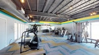 This fitness center offers a well-appointed workout environment designed to meet a variety of exercise needs. The space is defined by its striking industrial-style ceiling, where exposed steel trusses, corrugated metal, and visible ductwork create an open, airy atmosphere with generous overhead clearance. Pendant Edison-style bulbs and ceiling fans add both functional lighting and a contemporary aesthetic touch.

The main floor is laid out to maximize usability, featuring a diverse array of cardio equipment including stair climbers, treadmills, and stationary bikes. A dedicated strength training area provides a rack of free weights and resistance machines, catering to a full-body workout routine. Full-length mirrors line the walls, enhancing the sense of space while allowing users to monitor their form.

The flooring consists of colorful modular carpet tiles in shades of teal, yellow, and gray, which complement the vibrant horizontal accent stripes painted along the upper walls in matching tones. Together, these design elements create an energetic and motivating atmosphere. The single-level layout ensures easy accessibility throughout, making this fitness center a functional and visually dynamic space for exercise.