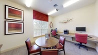 This single-floor business center and lounge offers a functional yet comfortable environment designed to accommodate both productivity and relaxed gathering. The space is divided into two distinct zones that flow naturally into one another.

The workstation area is anchored by a long granite-topped countertop desk that runs along the wall beneath a large window dressed with bold red Roman shades and horizontal blinds. The window overlooks a lush outdoor tree canopy, filling the area with natural light. A red executive chair, computer monitor, and a multifunction printer/scanner complete the fully equipped workspace. A decorative wall clock and a sculptural wire-and-branch wall accent add personality to the area.

Adjacent to the workstation is a casual meeting and lounge zone centered around a round wooden table with four red leather chairs, ideal for small group discussions or collaborative work. Comfortable striped armchairs in warm orange and red tones are tucked into a cozy seating nook, accented by a curated gallery wall of framed bird-motif canvas art pieces arranged in a dynamic cluster. Framed artwork with rich red tones adorns additional walls, contributing to the cohesive warm color palette throughout.

Carpeted flooring, recessed and fluorescent overhead lighting, and cream-toned walls create a clean, welcoming atmosphere throughout the space.