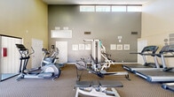 Located at 10139 Woodley Avenue in North Hills, CA, this fitness center is a compact yet thoughtfully outfitted facility designed to support a well-rounded workout routine. The space spans a single floor and is organized to maximize usability, with equipment arranged to allow comfortable movement throughout.

The facility features soaring vaulted ceilings that lend an open, airy feel to the room, preventing any sense of confinement despite its modest footprint. Natural light floods the interior through a row of clerestory windows positioned near the top of the walls, as well as through large sliding glass doors that provide direct access to an outdoor patio or courtyard area. The neutral-toned walls and patterned carpet flooring contribute to a clean, functional aesthetic.

Cardio equipment is well represented, with multiple treadmills, elliptical trainers, a stationary bike, and a rowing machine available for use. Strength training is supported by a multi-station cable machine, adjustable benches, and additional resistance equipment. A wall-mounted mirror enhances the sense of space and allows users to monitor their form. Safety essentials, including a fire extinguisher and hand sanitizer dispenser, are also present. An adjoining room provides additional utility to the overall facility.