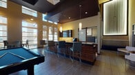 This contemporary amenity lounge is thoughtfully designed for socializing and recreation, offering a range of distinct activity zones within a single, open-concept floor. Soaring double-height ceilings with clerestory windows flood the space with natural light, creating an airy, upscale atmosphere throughout.

At the heart of the space sits a full-size billiards table set atop wood-look tile flooring with a subtle wave-patterned area rug beneath. Surrounding this focal point are high-top tables and bar stools, ideal for casual gatherings. A sophisticated wet bar anchors one side of the room, featuring rich wood cabinetry with glossy finishes, a light stone countertop, pendant lighting, and a coffee station—perfect for entertaining.

Adjacent to the bar, a dedicated lounge area offers a generous L-shaped leather sectional sofa paired with round ottomans, all oriented toward a large wall-mounted flat-screen television with under-cabinet accent lighting. A sliding barn door adds a modern architectural detail nearby.

A cozy nook with mosaic tile-clad banquette seating and abstract wall art provides an intimate retreat within the larger space. Throughout, warm gold and tan tones are complemented by deep espresso woodwork and dark ceiling accents, resulting in an elegant yet inviting environment suited for both relaxed lounging and lively social events.