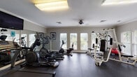 Located at 2400 Cluette Dr in Raleigh, NC, this dedicated fitness center offers a well-equipped, single-floor workout facility designed to support a full range of exercise routines. The space is finished with durable rubber tile flooring throughout, providing a safe and functional surface for all types of training activities.

The main workout area is generously sized and outfitted with a variety of cardio equipment, including treadmills, elliptical machines, and a recumbent bike. A multi-station cable and weight machine anchors the strength training section, complemented by a dumbbell rack and a flat workout bench for free weight exercises. Large mirrors line one wall, enhancing the sense of space and allowing users to monitor their form.

Crown molding and a neutral gray color palette give the space a polished, finished appearance. Ample natural light filters through multiple windows fitted with white shutters, while overhead fluorescent fixtures and a ceiling fan ensure consistent lighting and airflow. A wall-mounted television is positioned for visibility across the cardio area.

A secondary zone near the entrance features additional open floor space, seating, and access to an adjoining room through glass-paneled doors. Informational signage and sanitizing supplies are conveniently placed throughout. Overall, the facility presents a clean, organized, and welcoming environment suited for daily fitness use.