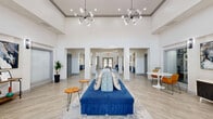 Located at 1533 Ellis Rd in Durham, NC, this single-floor community space offers a thoughtfully designed open-concept layout that seamlessly blends functionality with modern style. Soaring ceilings throughout create a sense of grandeur, while light wood-look flooring and crisp white walls lend a clean, contemporary aesthetic.

The entrance welcomes visitors with an expansive, airy foyer featuring herringbone-patterned flooring, bold abstract artwork, and direct sightlines into adjacent spaces — including a glass-enclosed fitness room stocked with equipment. Wall sconces and track lighting provide warm, layered illumination.

The living area is anchored by bold navy blue cabinetry with gold hardware beneath a shiplap accent wall, where open natural wood shelving adds warmth. This flows effortlessly into the kitchen, which features striking cobalt blue shaker-style cabinetry topped with white quartz countertops accented by dramatic veining. A subway tile backsplash, matte black fixtures, a deep undermount sink, and stainless steel appliances complete the polished culinary space.

A connecting hallway ties the zones together, maintaining an open and navigable floor plan. The overall ambiance is sophisticated yet inviting — a well-appointed gathering space that balances bold design choices with a welcoming, community-oriented atmosphere.