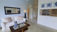 This single-floor residence features a spacious living area distinguished by notably high ceilings that lend an airy, open quality to the interior. The living room is finished with light-toned carpet underfoot and soft blue-gray walls, creating a calm, coastal ambiance. A large rectangular mirror with a bold black frame is mounted on one wall, visually expanding the space, while nautical-themed artwork adds a decorative accent nearby. A built-in niche on the opposite wall provides a display or storage feature that adds architectural interest.

The living room connects openly to the rest of the home through a wide, light-filled corridor leading toward a white panel door at the entry. Light tile flooring transitions the space from the carpeted living area into adjacent rooms. A pass-through opening to what appears to be a kitchen or dining area is visible along one wall, enhancing the open-concept feel and allowing for easy interaction between spaces. Natural light filters through the home, contributing to its bright, welcoming atmosphere. Overall, the space presents a clean, well-proportioned layout suited for comfortable everyday living.
