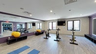 Quincy Yoga is a thoughtfully designed single-floor wellness studio that seamlessly blends functionality with a welcoming atmosphere. The main exercise room features light hardwood-style flooring that provides a clean, contemporary foundation throughout the space. A full-length wall mirror along one side visually expands the room while allowing practitioners to monitor their form during classes. The studio is equipped with a row of stationary cycling bikes, making it well-suited for spin and cardio-focused sessions, while yoga mats on the floor signal its versatility for yoga and stretching classes. Exercise balls and additional equipment are neatly stored along the mirrored wall. Natural light pours in through multiple windows fitted with dark frames, creating a bright and energizing environment, while a wall-mounted flat-screen television adds a modern touch and supports instructor-led programming. The walls feature a neutral textured finish accented by a bold purple accent wall, lending the studio a distinct and motivating character. Recessed ceiling lighting ensures the space remains well-lit at all times. The overall ambiance is clean, open, and purposefully arranged to support a variety of fitness and wellness activities.