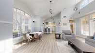 Located in Pleasanton, CA (94566), this single-floor community space combines thoughtful design with a clean, contemporary aesthetic. The interior is defined by soaring vaulted ceilings that lend an airy, expansive quality to the main open-plan area, which seamlessly integrates a lounge, dining zone, and kitchen into one cohesive layout.

The lounge area features comfortable gray upholstered seating arranged around a low coffee table atop a soft area rug, with large windows fitted with blinds that invite generous natural light while maintaining privacy. A glass-paned exterior door opens directly to an outdoor patio area, offering views of landscaped greenery and a pool beyond.

The dining area is anchored by a round white table surrounded by modern taupe chairs with gold-toned legs, positioned beneath an arched window that frames outdoor views and floods the space with daylight. Stylish globe pendant lights hang from the vaulted ceiling, complementing the wall-mounted sconces throughout.

The kitchen is efficiently designed with white cabinetry, a subway tile backsplash, light stone countertops, and stainless steel appliances including a full-size refrigerator. A peninsula with bar seating connects the kitchen to the main space. Light wood-look flooring runs throughout, tying each zone together under a cohesive soft blue-gray and white color palette that creates a calm, welcoming ambiance.
