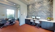This contemporary business center is thoughtfully designed to support productivity in a polished, professional environment. The focal point of the main workspace is a dramatic floor-to-ceiling accent wall clad in large-format geometric tiles with a bold chevron pattern in cool blue and gray tones, lending the space a distinctive, upscale aesthetic. Built-in dark wood cabinetry runs the length of this feature wall, supporting a clean white countertop workstation equipped with dual monitors, keyboards, and ample desk space for multiple users to work simultaneously. Ergonomic white and chrome task chairs complete the workstation setup, ensuring comfort during extended use. Recessed lighting in a coffered ceiling provides even, functional illumination throughout the space, while warm terracotta-toned carpet adds a welcoming contrast to the room's cool color palette. Adjacent to the main workspace, a glass-walled private room offers a more secluded setting, furnished with additional seating and benefiting from natural light through a nearby window. The transparent partition maintains visual connectivity while providing a quieter, enclosed area suitable for meetings or focused individual work. Together, the two areas create a well-rounded, amenity-rich workspace balancing open collaboration with private functionality.