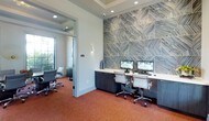 This contemporary business center is thoughtfully designed to support productivity in a polished, professional environment. The focal point of the main workspace is a dramatic floor-to-ceiling accent wall clad in large-format geometric tiles with a bold chevron pattern in cool blue and gray tones, lending the space a distinctive, upscale aesthetic. Built-in dark wood cabinetry runs the length of this feature wall, supporting a clean white countertop workstation equipped with dual monitors, keyboards, and ample desk space for multiple users to work simultaneously. Ergonomic white and chrome task chairs complete the workstation setup, ensuring comfort during extended use. Recessed lighting in a coffered ceiling provides even, functional illumination throughout the space, while warm terracotta-toned carpet adds a welcoming contrast to the room's cool color palette. Adjacent to the main workspace, a glass-walled private room offers a more secluded setting, furnished with additional seating and benefiting from natural light through a nearby window. The transparent partition maintains visual connectivity while providing a quieter, enclosed area suitable for meetings or focused individual work. Together, the two areas create a well-rounded, amenity-rich workspace balancing open collaboration with private functionality.
