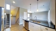 This three-floor residence offers a thoughtfully designed layout spread across multiple levels, connected by a staircase visible from the main living area. The kitchen serves as a central feature of the home, showcasing white shaker-style cabinetry topped with dark, glossy countertops that provide a striking contrast. A double stainless steel sink with a modern gooseneck faucet anchors the kitchen island, which also accommodates a stainless steel dishwasher beneath the counter. Two pendant lights hang above the island, adding focused task lighting and a contemporary aesthetic. The open layout allows the kitchen to flow seamlessly into an adjoining living or dining area, where a ceiling fan provides additional air circulation and ambient lighting. The space is finished with light hardwood-style flooring throughout, complementing the neutral gray walls that lend a clean, modern feel. Recessed lighting in the ceiling ensures the space remains well-lit. A white interior door leads to additional rooms, while a window allows natural light to filter in, brightening the overall atmosphere of this well-appointed, multi-story home.