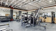 This fitness center offers a spacious, single-floor layout designed to accommodate a comprehensive range of workout activities. The open-truss ceiling, painted white and left exposed, creates an airy, industrial aesthetic while contributing to the sense of generous vertical space throughout the room. Large floor-to-ceiling windows line the perimeter, flooding the interior with natural light and providing views of the surrounding outdoor environment.

The flooring consists of durable, dark rubber gym mats well-suited for heavy equipment and high-traffic use. The space is equipped with an extensive selection of strength-training machines, including multi-station cable systems, a leg press, chest press, and lat pulldown units. A dedicated free weights area features a full rack of dumbbells alongside flat and adjustable benches. Cardio equipment includes multiple treadmills, an elliptical, and a stair climber, all positioned near the large windows to take advantage of natural light and outdoor views.

Accent walls combining dark vertical slat paneling and warm wood tones add a modern, polished feel to the space. Full-length mirrors are strategically placed to visually expand the room. A wall-mounted water fountain and a wall-mounted television are also present. The overall ambiance is clean, functional, and professional, making this fitness center a well-rounded facility for all levels of training.