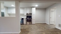 This single-floor unit offers a thoughtfully designed layout with a clean, neutral aesthetic carried throughout each room. The kitchen is a standout feature, outfitted with crisp white shaker-style cabinetry, white countertops, and stainless steel appliances including a range, dishwasher, and refrigerator. A pass-through window connects the kitchen to the adjacent living area, creating an open and communicative flow between spaces. Wood-look flooring runs throughout the main living areas, adding warmth and a sense of continuity.

The living area is generously sized and anchored by a classic fireplace framed in deep navy, complemented by a ceiling fan overhead — a practical and inviting centerpiece for the room. Natural light enters through blinded windows, keeping the space bright while maintaining privacy.

The bedroom features plush wall-to-wall carpeting underfoot and a window that allows for natural light, creating a comfortable and quiet retreat. The bathroom is finished with white subway tile surrounding a full soaking tub and shower combination, with chrome fixtures and wood-look flooring adding a clean, polished look. The entryway provides a welcoming transition into the home, with a front door flanked by a window and direct sightlines into the kitchen area.