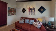 This single-floor unit is distinguished by its bold, jazz-inspired interior aesthetic carried consistently throughout every room. The living area features striking exposed dark wood beams across vaulted ceilings, carpet flooring, and large windows that allow natural greenery views to filter in, creating a warm and inviting atmosphere. Bold canvas artwork depicting jazz musicians anchors the walls, complemented by decorative diamond-shaped wall pieces and a rich burgundy sofa. The front entry opens directly into this living space, which connects to the adjoining kitchen.

The galley-style kitchen features light wood cabinetry, tile flooring, a double sink, a dishwasher, and an electric range. The layout is efficient and functional, with ample counter and cabinet storage along both walls.

The bedroom continues the music-themed decor with jazz club canvas prints and decorative crown wall art. It features carpeted floors, a textured ceiling, and dark wood furniture. The room offers enough space for a full-size bed and dresser.

The full bathroom includes a single-sink vanity with medium-toned wood cabinetry, a large mirror with overhead lighting, an above-toilet storage cabinet, and a tub-shower combination, providing practical and organized storage throughout.