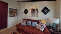 This single-floor unit is distinguished by its bold, jazz-inspired interior aesthetic carried consistently throughout every room. The living area features striking exposed dark wood beams across vaulted ceilings, carpet flooring, and large windows that allow natural greenery views to filter in, creating a warm and inviting atmosphere. Bold canvas artwork depicting jazz musicians anchors the walls, complemented by decorative diamond-shaped wall pieces and a rich burgundy sofa. The front entry opens directly into this living space, which connects to the adjoining kitchen.

The galley-style kitchen features light wood cabinetry, tile flooring, a double sink, a dishwasher, and an electric range. The layout is efficient and functional, with ample counter and cabinet storage along both walls.

The bedroom continues the music-themed decor with jazz club canvas prints and decorative crown wall art. It features carpeted floors, a textured ceiling, and dark wood furniture. The room offers enough space for a full-size bed and dresser.

The full bathroom includes a single-sink vanity with medium-toned wood cabinetry, a large mirror with overhead lighting, an above-toilet storage cabinet, and a tub-shower combination, providing practical and organized storage throughout.