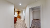 This contemporary single-floor residence offers a thoughtfully designed layout with clean lines and modern finishes throughout. The galley-style kitchen is a standout feature, showcasing warm wood cabinetry, stainless steel appliances including a professional-grade gas range with hood, built-in microwave and dishwasher, tile backsplash, and sleek countertops — all set against light hardwood-style flooring that flows seamlessly through the entire unit. The open living and dining areas are bright and airy, bathed in natural light from expansive floor-to-ceiling windows and sliding glass doors that frame sweeping city views. Sheer vertical blinds provide adjustable privacy while maintaining the sense of openness. A curved architectural column adds a refined design detail to the corner window area. The bedroom features generous proportions, direct access to a walk-in closet, and continued floor-to-ceiling window exposure with outdoor views. The bathroom is finished with a white quartz vanity, large wall mirror, wood-toned cabinetry, and a combined tub and shower enclosure with glass doors. Elegant pendant lighting in the living area and recessed lighting throughout complete the polished, contemporary aesthetic of this well-appointed residence.
