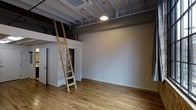 This loft-style space makes an immediate impression with its soaring ceilings that reveal exposed structural beams, ductwork, and industrial piping — a hallmark of converted warehouse or urban loft architecture. The open layout is anchored by warm, natural hardwood floors that run throughout, lending a welcoming contrast to the raw, industrial overhead elements.

A standout feature is the elevated mezzanine level, accessible via a wooden ladder with a natural finish. The mezzanine platform adds a versatile upper-level area, ideal for use as a sleeping loft, storage space, or a private retreat, while a set of white panel doors below the loft platform provides additional enclosed storage or closet space.

The main living area is expansive and open, with clean, light-gray painted walls that create a neutral, versatile backdrop. Overhead, a single pendant light fixture provides focused ambient illumination, complementing the industrial ceiling aesthetic. The ceiling-mounted HVAC unit ensures efficient climate control while maintaining the open, uncluttered feel of the space.

The overall ambiance is a refined blend of industrial and modern sensibilities — raw structural elements balanced by clean finishes and warm wood tones — making this a highly adaptable and characterful space.