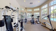 This single-floor fitness center is thoughtfully designed to provide a bright and motivating workout environment. The space features a curved bank of large, multi-pane windows that flood the room with natural light and offer scenic views of a sparkling pool and surrounding tropical landscaping, including mature palm trees. The warm, neutral-toned walls and carpeted flooring contribute to a clean and welcoming atmosphere.

The gym is well-appointed with a comprehensive selection of equipment to support both cardiovascular and strength training workouts. Cardio offerings include two treadmills, an elliptical trainer, a stationary upright bike, and a stair-stepper, with several machines branded by TRUE Fitness. Strength training is supported by a multi-station cable machine with a bench attachment, as well as additional resistance equipment. A wall-mounted flat-screen television is positioned above the treadmill area for entertainment during workouts.

The facility benefits from excellent ventilation with ceiling vents, recessed lighting, and transom windows that enhance airflow and brightness. A glass-paneled service window, waste receptacles, and informational postings along the walls add functional touches. Glass doors on multiple sides provide easy access to the outdoor surroundings, seamlessly connecting the indoor fitness experience with the resort-like exterior environment.