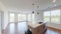 This single-floor residence offers a thoughtfully designed open-concept layout that seamlessly connects the kitchen, dining, and living areas. The kitchen serves as a central hub, featuring a large island with an undermount sink, light quartz countertops, and wood-toned cabinetry with sleek hardware. Stainless steel appliances, including a French door refrigerator, electric range, built-in microwave, and dishwasher, are complemented by a white subway tile backsplash. Black pendant lights hang above the island, adding a contemporary accent.

The expansive living area is bathed in natural light from a generous bank of windows that frame green outdoor views, with a ceiling fan providing additional comfort. Hardwood-style flooring runs throughout the main living spaces, while carpeting is found in the bedrooms, which are bright and well-proportioned with large windows.

Two bathrooms serve the unit: one featuring a glass-enclosed walk-in shower, dual-sink vanity with wood-toned cabinetry, and hardwood-style flooring, while the second offers a tub/shower combination with a matching vanity. The overall aesthetic is clean, modern, and light-filled throughout.