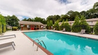 Located at 2050 Settle Cir SE in Atlanta, Georgia, this outdoor amenity area centers around a generously sized rectangular swimming pool with vivid turquoise water and classic brick coping. The pool deck is finished in smooth concrete, offering ample space for lounging, and is furnished with multiple chaise lounge chairs positioned along the pool's perimeter for relaxation and sunbathing.

A standout feature of the space is a bold, dark-painted pergola structure anchored by stone pillars on one side of the deck. Beneath and adjacent to it sits an outdoor grill station alongside patio seating with a table and umbrella, creating a well-equipped outdoor entertaining area ideal for gatherings.

A dedicated pool house with a red-accented door is situated at one end of the deck, providing a functional support structure for the area. The entire pool zone is enclosed by a black metal security fence, ensuring safety while maintaining an open, airy feel.

The surroundings are richly landscaped with tall evergreen trees, mature hardwoods, and manicured shrubs that provide natural privacy and a lush green backdrop. The combination of the vibrant pool, shaded pergola, and verdant landscaping creates a serene and inviting outdoor retreat within the heart of Atlanta's East Side neighborhood.