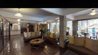 This single-floor space exudes a distinctive character rooted in rustic, Southwestern-inspired design. Rich, dark terracotta tile floors run throughout, lending warmth and visual continuity across the entire layout. Exposed brick columns punctuate the open interior, reinforcing the space's structural character and adding an organic, textured element to the overall aesthetic.

A glass-and-dark-wood partitioned enclosure houses a private office or reception area, offering transparency while maintaining a degree of separation from the main open areas. Pendant lighting with warm-toned globes hangs at intervals, complementing recessed ceiling fixtures to create a layered, inviting ambiance.

The seating area is furnished with substantial wooden sofas and armchairs adorned with patterned cushions in earthy gold and brown tones, arranged around a carved wooden coffee table. Large sliding glass doors and plantation-shuttered windows allow generous natural light to filter in, with views of lush outdoor greenery and a shaded patio beyond.

Additional features include a wall-mounted flat-screen television, a decorative console table with plants and accessories, and bold double entry doors finished in dark lacquer, contributing to the space's layered, character-rich presentation.