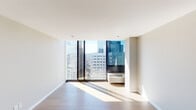 This single-floor residence offers a thoughtfully designed layout with a clean, contemporary aesthetic throughout. The living area is a standout feature, characterized by expansive floor-to-ceiling windows framed in dark metal that frame sweeping views of a dense urban skyline, flooding the space with abundant natural light. Wide-plank hardwood flooring in a warm tone runs throughout the main living areas, contributing to a cohesive and polished look.

The kitchen is designed with a galley-style configuration, featuring dark cabinetry that extends to the ceiling, stainless steel appliances including a refrigerator, range, and microwave, white countertops, and a grey subway tile backsplash that adds a modern industrial touch.

The bedroom provides a generous, well-proportioned space with warm-toned hardwood flooring, neutral grey walls, and a closet for storage. Notably, an in-unit stacked washer and dryer is conveniently housed within a dedicated alcove in the bedroom area.

The bathroom is finished with marble-style wall tiles surrounding a full soaking tub and shower combination, complemented by brushed nickel fixtures and a vessel sink, offering a clean and refined aesthetic throughout.