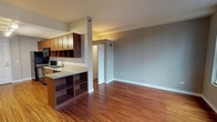 Situated in the heart of Chicago's Loop at 60 E Randolph Street, this single-floor residence offers a well-designed open-concept layout with a cohesive blend of modern finishes and warm tones throughout. Rich hardwood flooring runs continuously across the main living areas, lending warmth and a sense of continuity to the space.

The kitchen is thoughtfully appointed with warm-toned wood cabinetry, granite countertops, a stainless steel sink, and a full suite of black appliances including an electric range, over-the-range microwave, and dishwasher. A peninsula with built-in open shelving connects the kitchen to the living area, providing additional storage and serving as a natural division between the two spaces.

The open living area is generously proportioned and bathed in natural light from large double-hung windows that frame impressive views of the surrounding Chicago skyline — a notable feature that adds a dynamic urban backdrop to the interior.

The bathroom is clean and functional, featuring white tile throughout, a soaking tub with shower, white tile surround, and a modern vessel sink set atop a wood vanity with a framed mirror. The overall design palette of neutral grays and whites creates a bright, fresh atmosphere. This well-located urban residence offers practical amenities within one of Chicago's most central and vibrant neighborhoods.
