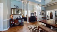 Located at 3919 Carnegie Ave in Myrtle Beach, SC, this single-floor space exudes a warm, upscale ambiance rooted in classic Southern design. The main gathering area impresses with soaring vaulted ceilings adorned with decorative crown molding and elevated display shelving lined with tropical greenery and artwork. Rich hardwood flooring flows throughout, complemented by patterned area rugs that define the various seating zones.

The space is generously appointed with eclectic, elegant furnishings — including leather arm chairs, ornate carved wood pieces, and upholstered accent seating — all arranged to encourage both conversation and comfort. A tiled decorative fireplace with a detailed mantle serves as a striking focal point, flanked by French doors that flood the interior with abundant natural light and provide views of the outdoor surroundings.

Structural white columns add architectural interest throughout the open layout, while wrought iron wall accents and framed artwork lend a refined, curated character. A dedicated coffee station, outfitted with a coffee maker and accessory display, offers a welcoming amenity for visitors. A built-in dark wood display shelf near the entrance showcases decorative objects and greenery, contributing to the space's inviting, resort-inspired atmosphere.