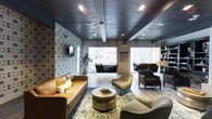 This single-floor space presents a thoughtfully designed lounge area that blends industrial and contemporary aesthetics. The dark, exposed ceiling features recessed lighting and ceiling fans, lending a moody yet polished ambiance to the interior. A striking geometric patterned accent wall anchors one side of the room, providing a bold visual contrast against the softer furnishings throughout the space.

The seating arrangement is diverse and intentional, centering around a cylindrical metallic coffee table paired with a tan leather sofa and complementary lounge chairs upholstered in neutral tones. Area rugs with subtle, ornate patterns define the seating zones and add warmth underfoot. One of the most distinctive features of the space is the pair of suspended swing chairs hanging from the ceiling near large windows, which flood the area with natural light and offer views of the surrounding urban landscape.

Toward one side of the room, a more intimate seating cluster with classic tufted armchairs provides an alternative gathering spot. Open shelving units along the wall add functional storage and display space. Together, these elements create a versatile, well-appointed lounge suited for both relaxation and social interaction.