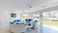 This single-floor lounge space exudes a fresh, coastal aesthetic defined by a crisp white palette and vibrant blue accents throughout. The main gathering area is generously proportioned, featuring curved white wicker sofas and chairs adorned with an array of sky-blue and navy throw pillows, all arranged around a decorative white coffee table. The open, airy atmosphere is enhanced by large windows that flood the room with abundant natural light and offer views of the surrounding greenery and outdoor amenities. Ceiling fans overhead provide comfortable air circulation, adding to the relaxed ambiance of the space. A wall-mounted flat-screen television makes the lounge well-suited for both casual socializing and organized gatherings. Glass doors on one side of the room open directly onto an outdoor patio area, where patio furniture and teal market umbrellas are visible alongside a well-maintained lawn, seamlessly blending indoor and outdoor living. The smooth, light-toned flooring and white walls throughout keep the space feeling clean, modern, and inviting. Overall, this lounge delivers a resort-inspired atmosphere ideal for relaxation and social gatherings.