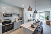 Located in Fort Myers, FL, this well-appointed 2-bedroom, 2-bathroom residence offers a thoughtfully designed single-floor layout that balances comfort and modern style. The open-concept living area seamlessly connects the kitchen, dining, and living spaces, all bathed in natural light from large sliding glass doors leading to a private balcony with pleasant outdoor views. The kitchen is a standout feature, boasting a generous quartz-topped island with bar seating, sleek gray cabinetry, stainless steel appliances, and stylish pendant lighting overhead. Light wood-tone flooring flows throughout the main living areas, enhancing the bright, airy ambiance. The combined dining and living room provides a comfortable gathering space, while a ceiling fan adds to the breezy Florida feel. Both bedrooms are carpeted and well-sized, with the primary bedroom offering ample natural light and a spacious walk-in closet with built-in shelving. The two bathrooms are clean and modern — one featuring a tub/shower combination and the other a glass-enclosed shower. Additional highlights include a laundry area, utility room, hallway, and five closets throughout, ensuring exceptional storage. This residence offers a polished, contemporary lifestyle in a practical and efficient floor plan.