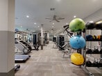 Located at 816 Addicks Howell Rd in Houston, TX, this fitness center offers a comprehensive workout facility across a generously sized, single-floor layout. The space is thoughtfully designed with a neutral color palette — warm beige walls accented by a two-tone gray lower section — and durable, light-toned carpet tile flooring throughout that gives the space a clean, modern aesthetic.

The facility is divided into distinct workout zones. The cardio area features a row of treadmills, elliptical trainers, stationary cycling bikes, and rowing machines, several of which are positioned near large floor-to-ceiling windows that overlook a landscaped courtyard, filling the space with natural light. Ceiling fans are installed throughout to promote air circulation during workouts.

The strength training section is equally well-appointed, housing a variety of cable and selectorized machines, a power rack with an organized barbell and weight storage system, and a full rack of dumbbells. Stability balls and medicine balls of varying sizes are neatly displayed on a dedicated rack, supporting functional and dynamic training needs.

Full-length mirrors along key walls enhance the sense of openness and allow users to monitor their form. Recessed lighting illuminates the entire space evenly, contributing to a bright and inviting atmosphere. The facility also includes a glass-partitioned area, adding architectural distinction to the overall design.