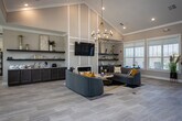 The Sunrise Ridge clubhouse at 2401 Sunrise Ridge Way in Knoxville, Tennessee, is a thoughtfully designed single-story community facility that combines comfort, functionality, and modern aesthetics. The main lounge serves as the heart of the space, featuring soaring vaulted ceilings, light wood-look tile flooring, and an elegant gold chandelier that anchors the room. A built-in fireplace with a herringbone tile surround and a wall-mounted television creates a welcoming focal point, while plush grey sofas accented with mustard yellow pillows add a contemporary flair. Large double-glass entry doors and expansive windows flood the interior with natural light and offer views of the surrounding outdoors. A stylish credenza with floating shelves provides decorative display space, seamlessly connecting the lounge to additional seating and dining areas. The catering kitchen is well-appointed with granite countertops, dark cabinetry, a striking geometric tile backsplash, pendant lighting, and a central island — ideal for community gatherings. A dedicated fitness center rounds out the amenities, equipped with cardio machines including treadmills and stationary bikes, strength equipment, free weights, a mirrored wall, and durable rubber flooring, all set beneath high vaulted ceilings with ample natural light from multiple windows.