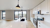This contemporary single-floor residence offers a thoughtfully designed open-concept layout that seamlessly connects the kitchen and living areas. The kitchen is a standout feature, showcasing a large island with a striking two-tone design — a crisp white quartz countertop paired with a warm wood base — and an undermount black sink accented by matching matte black pendant lights overhead. White shaker-style cabinetry extends to the ceiling, providing generous storage, while a classic subway tile backsplash adds timeless texture. Stainless steel appliances, including a range, over-the-range microwave, and refrigerator, complete the well-appointed cooking space. Light wood-tone flooring flows throughout the unit, enhancing the bright and airy atmosphere. The open living area is bathed in natural light from floor-to-ceiling black-framed windows and a full-width sliding glass door that leads to a private outdoor balcony with open neighborhood views. The bedroom offers a clean, neutral palette with recessed lighting and a sliding closet door for convenient storage. The bathroom features a walk-in shower, tile flooring, and a minimalist design. Overall, the space presents a polished, modern aesthetic with a cohesive neutral color scheme.
