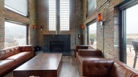 This striking industrial-style lounge space masterfully blends raw architectural character with refined comfort. Exposed brick walls rise dramatically to towering ceiling heights, creating an expansive, loft-like atmosphere throughout. Visible pipework and conduit run along the walls and ceiling, celebrating the building's utilitarian heritage while adding to its authentic industrial aesthetic.

The main lounge area is anchored by a bold, dark granite fireplace flanked by floor-to-ceiling glass block windows that flood the space with diffused natural light. Tufted brown leather sofas and sectionals are arranged around substantial stone-topped coffee tables, creating an inviting and upscale seating environment. Wall-mounted art pieces and warm amber sconces add curated style against the textured brick backdrop.

A built-in bar with counter seating provides a dedicated service area, while a large wall-mounted display screen near the stairwell landing offers media capabilities suited for events or presentations. Metal mesh railings define the stairwell access point, contributing to the cohesive industrial design language.

Large windows frame views of the surrounding cityscape, and a set of glass doors opens directly onto a spacious outdoor rooftop deck furnished with rattan seating and dining tables — an exceptional extension of the interior that invites guests to enjoy open-air urban views.