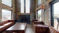 This striking industrial-style lounge space masterfully blends raw architectural character with refined comfort. Exposed brick walls rise dramatically to towering ceiling heights, creating an expansive, loft-like atmosphere throughout. Visible pipework and conduit run along the walls and ceiling, celebrating the building's utilitarian heritage while adding to its authentic industrial aesthetic.

The main lounge area is anchored by a bold, dark granite fireplace flanked by floor-to-ceiling glass block windows that flood the space with diffused natural light. Tufted brown leather sofas and sectionals are arranged around substantial stone-topped coffee tables, creating an inviting and upscale seating environment. Wall-mounted art pieces and warm amber sconces add curated style against the textured brick backdrop.

A built-in bar with counter seating provides a dedicated service area, while a large wall-mounted display screen near the stairwell landing offers media capabilities suited for events or presentations. Metal mesh railings define the stairwell access point, contributing to the cohesive industrial design language.

Large windows frame views of the surrounding cityscape, and a set of glass doors opens directly onto a spacious outdoor rooftop deck furnished with rattan seating and dining tables — an exceptional extension of the interior that invites guests to enjoy open-air urban views.