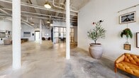 Located in Augusta, Georgia, this expansive single-floor lobby blends industrial character with contemporary design to create a polished and functional commercial space. Polished concrete floors stretch throughout the open layout, reflecting light and reinforcing a clean, modern aesthetic. Soaring ceilings with exposed structural beams, ductwork, and pendant lighting give the space an authentic industrial feel while maintaining an inviting atmosphere.

The lobby is organized into several distinct zones. A lounge area features a curved cognac velvet sofa set atop a diamond-patterned area rug, anchored by a dramatic globe chandelier and a built-in green shelving unit that adds warmth and color. Nearby, a second seating area showcases a curated gallery wall of mixed mirrors and framed art above a coordinated sofa and chairs arrangement. A pair of tufted armchairs near an arched doorway with whitewashed brick walls offers a more intimate seating nook.

A glass-walled conference room enclosed by sleek black steel-framed partitions provides a private meeting space while maintaining visual connectivity to the open floor. Structural white columns punctuate the layout, adding architectural rhythm. The combination of warm lighting, thoughtful furnishings, and industrial materials creates a sophisticated yet approachable environment well-suited for a professional or creative workplace setting.