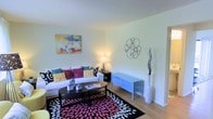 This two-floor space offers a well-lit and inviting living area that serves as a central gathering point. The living room features light hardwood flooring that runs throughout, complementing the crisp white walls and contributing to an open, airy feel. Natural light enhances the brightness of the room, highlighting the clean, neutral palette of the walls and ceiling.

The living area is thoughtfully arranged, with ample space for comfortable seating. A white sofa dressed with colorful accent pillows in teal, pink, and gold anchors the room, paired with a sleek black oval coffee table resting atop a bold black, white, and pink patterned area rug that adds a vibrant focal point to the space. A light blue media console provides functional storage and display space along one wall, while decorative wall art and a sculptural metallic wall accent contribute personality and visual interest. A tall white floor vase with branching décor adds vertical dimension to the corner.

The overall ambiance is bright and modern, with a playful use of color against a neutral backdrop, creating a space that feels both lively and comfortable across its two floors.