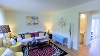This two-floor space offers a well-lit and inviting living area that serves as a central gathering point. The living room features light hardwood flooring that runs throughout, complementing the crisp white walls and contributing to an open, airy feel. Natural light enhances the brightness of the room, highlighting the clean, neutral palette of the walls and ceiling.

The living area is thoughtfully arranged, with ample space for comfortable seating. A white sofa dressed with colorful accent pillows in teal, pink, and gold anchors the room, paired with a sleek black oval coffee table resting atop a bold black, white, and pink patterned area rug that adds a vibrant focal point to the space. A light blue media console provides functional storage and display space along one wall, while decorative wall art and a sculptural metallic wall accent contribute personality and visual interest. A tall white floor vase with branching décor adds vertical dimension to the corner.

The overall ambiance is bright and modern, with a playful use of color against a neutral backdrop, creating a space that feels both lively and comfortable across its two floors.