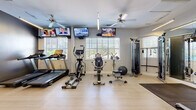 Located at 100 Madison Place Cir in Kernersville, NC, this fitness center offers a well-equipped, single-floor workout facility designed to support a comprehensive exercise routine. The space features light hardwood-style flooring throughout, neutral gray walls, and ample natural light streaming in through multiple white-shuttered windows, creating a bright and energizing atmosphere. Ceiling fans are positioned throughout the room to maintain air circulation during workouts.

The cardio section is equipped with treadmills, elliptical trainers, a recumbent bike, and spin bikes, providing a variety of options for cardiovascular exercise. Multiple wall-mounted flat-screen TVs are installed at strategic viewing angles, allowing users to stay entertained during workouts. A dedicated strength training area includes a cable machine, a functional trainer, and a weight bench, complemented by a dumbbell rack with a full range of free weights and a medicine ball station along the wall.

A large wall mirror enhances the sense of space and supports proper form during strength exercises. The facility also includes a rubber floor mat in the weight area for safety and equipment protection. The clean, organized layout and dual-access doors make the space practical and easy to navigate, offering residents a convenient and fully functional fitness amenity.