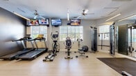 Located at 100 Madison Place Cir in Kernersville, NC, this fitness center offers a well-equipped, single-floor workout facility designed to support a comprehensive exercise routine. The space features light hardwood-style flooring throughout, neutral gray walls, and ample natural light streaming in through multiple white-shuttered windows, creating a bright and energizing atmosphere. Ceiling fans are positioned throughout the room to maintain air circulation during workouts.

The cardio section is equipped with treadmills, elliptical trainers, a recumbent bike, and spin bikes, providing a variety of options for cardiovascular exercise. Multiple wall-mounted flat-screen TVs are installed at strategic viewing angles, allowing users to stay entertained during workouts. A dedicated strength training area includes a cable machine, a functional trainer, and a weight bench, complemented by a dumbbell rack with a full range of free weights and a medicine ball station along the wall.

A large wall mirror enhances the sense of space and supports proper form during strength exercises. The facility also includes a rubber floor mat in the weight area for safety and equipment protection. The clean, organized layout and dual-access doors make the space practical and easy to navigate, offering residents a convenient and fully functional fitness amenity.