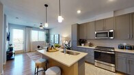 This thoughtfully designed single-floor residence combines contemporary style with functional living. The open-concept layout connects the kitchen and living area seamlessly, creating an inviting, airy atmosphere filled with natural light from large windows dressed with sheer curtains and blinds.

The kitchen is a standout feature, equipped with sleek grey shaker-style cabinetry, a herringbone tile backsplash, stainless steel appliances including a gas range, microwave, and French door refrigerator, and a generous island with a white quartz countertop and built-in sink. Pendant lighting above the island adds a refined touch, while wood-look flooring ties the space together.

The living room offers a comfortable retreat with a ceiling fan, access to an outdoor area through a glass door, and views to the outdoors. Two bedrooms provide restful spaces, each featuring carpeted floors, ample natural light, and window treatments for privacy. The primary bedroom is accented by a bold blue feature wall and room for a large bed.

The bathroom is well-appointed with grey cabinetry, a single undermount sink, and direct access to a spacious walk-in closet with extensive shelving and hanging storage. Throughout the residence, a neutral, modern palette and consistent finishes create a cohesive and polished living environment.