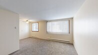 This 2-bedroom, 1-bathroom unit offers a practical and efficiently designed single-floor layout. The spacious primary bedroom is the largest room in the unit, featuring plush carpeting, neutral cream-toned walls, a ceiling fan with light fixture, baseboard heating, and two windows — including a large picture window with vertical blinds that allows for generous natural light. The second bedroom is more compact, also carpeted and neutrally finished, with a window offering outdoor views and its own baseboard heater. The galley-style kitchen is efficiently laid out with gray cabinetry lining both walls, a double-basin sink, laminate countertops, a coil-element electric range, and a refrigerator. The tile flooring in a warm earth-tone pattern adds a classic touch. The bathroom features retro-style tiling with a distinctive turquoise accent border, a full soaking tub with shower, and a wood-vanity with an integrated sink and mirror above. A connecting hallway ties the rooms together, completing the functional flow of the unit. Baseboard heating is present throughout, ensuring consistent warmth, and the overall color palette of warm beiges and whites creates a clean, neutral ambiance.