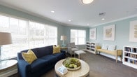 This single-story space features a well-appointed living area designed with a calm, welcoming atmosphere. The walls are painted in a soft blue-green hue, contributing to a serene and airy feel throughout. Recessed lighting in the white ceiling is complemented by a flush-mount overhead fixture, ensuring the space is well-lit at all times.

Large windows fitted with white horizontal blinds line the curved wall, allowing generous amounts of natural light to filter in while maintaining privacy. The flooring is covered in a neutral-toned carpet that grounds the room's coastal-inspired color palette.

The seating arrangement centers around a deep navy blue sofa accented with mustard yellow throw pillows, paired with a light gray armchair and matching ottoman. A round wooden coffee table serves as a focal point, styled with a decorative floral centerpiece and reading material. A built-in white bookshelf along one wall provides both storage and display space, while framed artwork adds visual interest. A yellow accent chair and a glass-base table lamp with a classic shade round out the thoughtfully curated furnishings, creating a balanced and inviting environment.