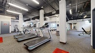 Located at 929 Jefferson St in Kansas City, Missouri, this fitness facility offers a thoughtfully designed, two-zone gym experience spread across a single floor. The space blends industrial and modern aesthetics, with exposed black steel beams, open ductwork, and painted ceilings in the main workout area contrasting with clean white walls and structural columns. Patterned carpet tile flooring runs throughout, providing both durability and a contemporary look.

The main gym area is generously proportioned, featuring a row of treadmills and cardio machines positioned alongside large wall mirrors that enhance the sense of openness. Accent red floor markers add a dynamic visual element, while natural light filters in through windows along the perimeter. A dedicated strength training zone includes adjustable weight benches and a cable machine, offering a comprehensive free weights and resistance training setup. Exposed brick accents add a nod to the building's historic character.

A separate cycling studio provides an intimate, focused environment for spinning classes. This room features a dropped tile ceiling with recessed lighting, a wall-mounted flat-screen television, and a mirrored wall, all contributing to an energizing yet contained atmosphere. The studio is accessed via glass-paneled sliding doors, maintaining visual connectivity with the main floor while providing acoustic separation. Together, the two spaces form a complete, well-appointed fitness destination.