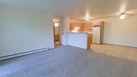 This single-floor residential space presents a clean, functional layout centered around an open-concept living and kitchen area. The living room is finished with wall-to-wall carpeting in a neutral tone, creating a comfortable and inviting atmosphere. A baseboard heating unit runs along the exterior wall, ensuring efficient warmth throughout the space. The kitchen is defined by a half-wall peninsula that separates it from the living area, providing both a visual boundary and additional counter space. Light wood cabinetry lines the kitchen walls, offering generous storage, while a full-size refrigerator and under-cabinet range hood complete the appliance setup. The flooring transitions from carpet in the living area to a hard surface in the kitchen, a practical detail that aids in maintenance. A hallway visible from the main living space leads to an additional room, which appears to include a bathroom with a window that allows natural light to filter through. The overall palette of the space is neutral and understated, with white walls and light-toned finishes throughout, lending the unit a bright and airy feel. The straightforward, efficient floor plan makes excellent use of the available space.