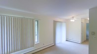 This single-floor space presents a clean, minimalist living area characterized by its bright, airy atmosphere. The room is finished with neutral cream-colored walls and light carpeting, creating a soft, cohesive palette throughout. Two windows dressed with vertical blinds allow natural light to filter into the space, enhancing its open feel. A larger window treatment, likely covering a sliding or full-length door, provides additional brightness and potential outdoor access. A ceiling fan with a light fixture is centrally positioned, offering both air circulation and ambient lighting. Baseboard heating units run along the lower walls, providing an efficient heating solution integrated seamlessly into the room's perimeter. The space is currently vacant, allowing for a clear view of the room's layout and architectural simplicity. The overall ambiance is calm and functional, with a neutral design that offers flexibility for a variety of furnishing styles. The single-floor layout provides convenient, step-free living throughout the entire space.