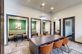 Located at 698 Basilica Bay Drive in Spring, TX, this thoughtfully designed communal space offers a versatile and professional environment for meetings, collaboration, and dining. The single-floor layout is efficiently organized to maximize functionality without sacrificing style.

At the heart of the space sits a large, dark-toned rectangular conference table surrounded by cognac-orange mid-century modern chairs, illuminated by a pair of contemporary white dome pendant lights. The patterned, multi-tone carpeting adds texture and warmth underfoot throughout the room.

A striking vertical wood slat partition serves as an architectural divider, adding a warm, organic contrast to the otherwise clean white walls and dark-framed doorways. Natural light filters in through tall windows dressed with dark drape panels, complemented by a seating nook furnished with neutral-toned lounge chairs.

Two semi-private dining booths flank the main area, each featuring bold green accent walls, vibrant artwork, and bench-style seating paired with sleek black chairs — ideal for smaller gatherings or casual meetings. A dedicated conference room, accessible via a frosted glass door, provides an enclosed space for private discussions. The overall ambiance is modern and inviting, balancing professional utility with contemporary residential warmth.